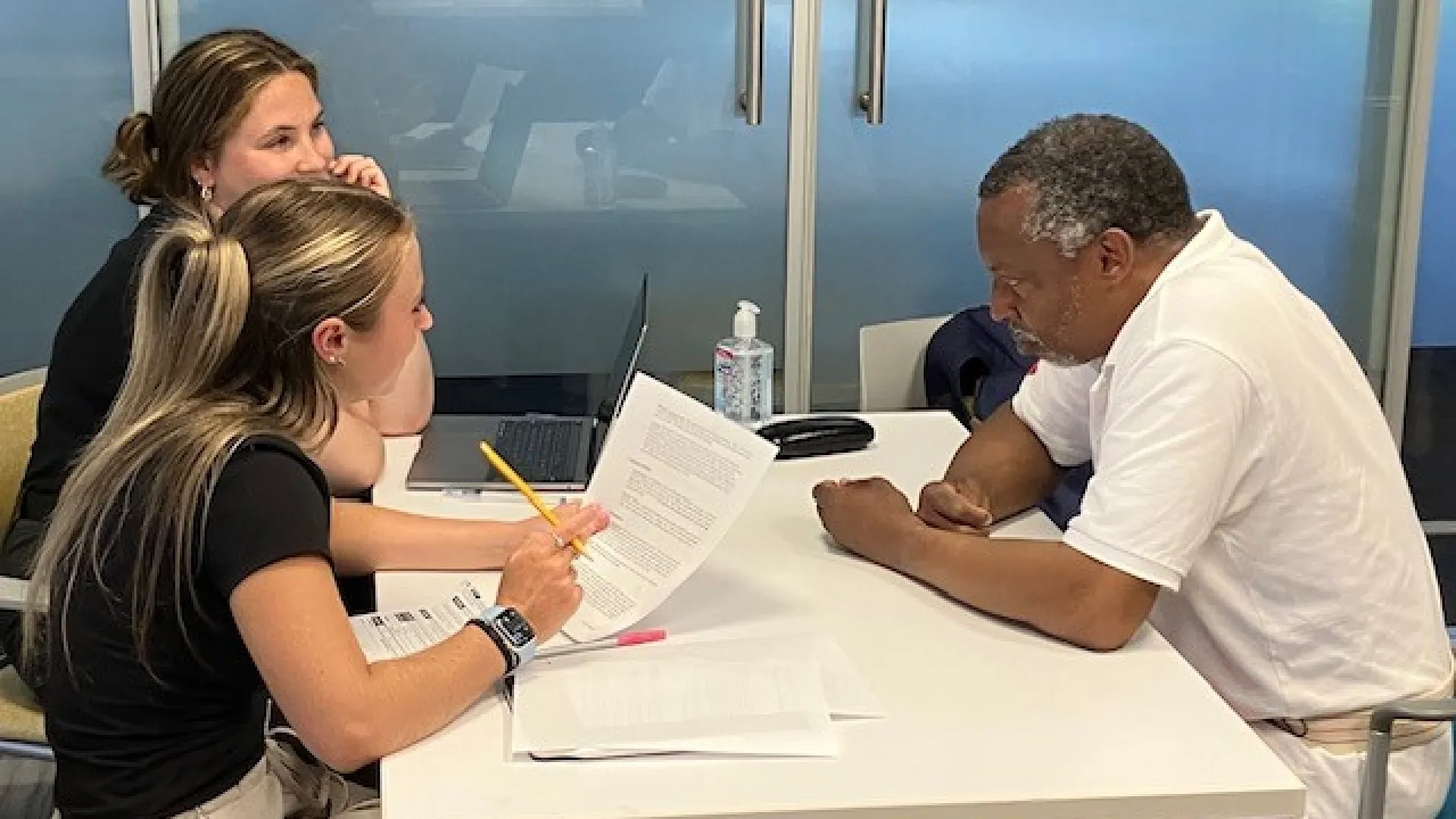 A man sits at a table while two women on the other side look at a computer and hold a paper