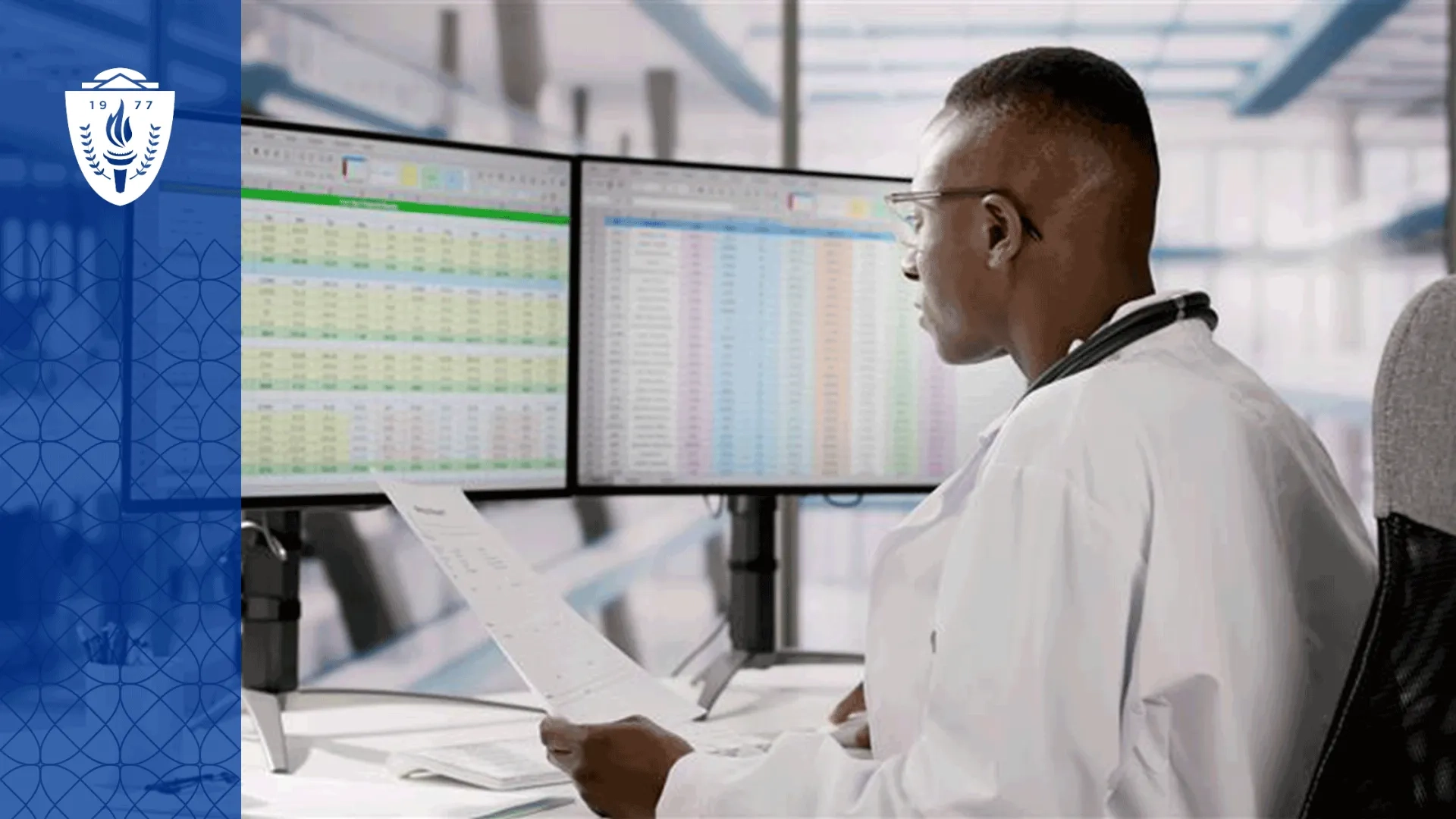 Man wearing lab coat sitting a desk and looking at spreadsheets on two computer monitors