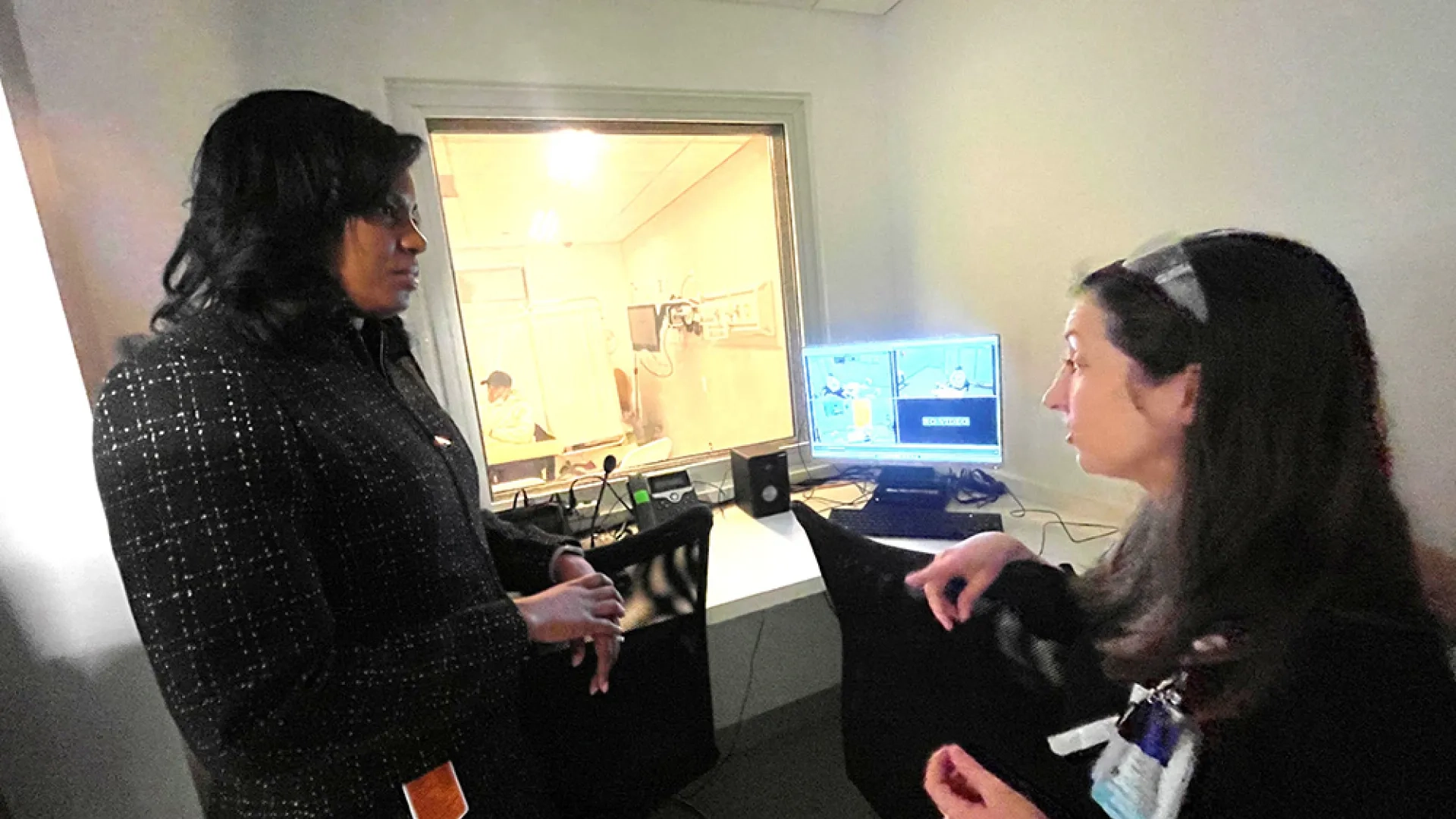 Two women talk to each other with computer equipment and a window in the background