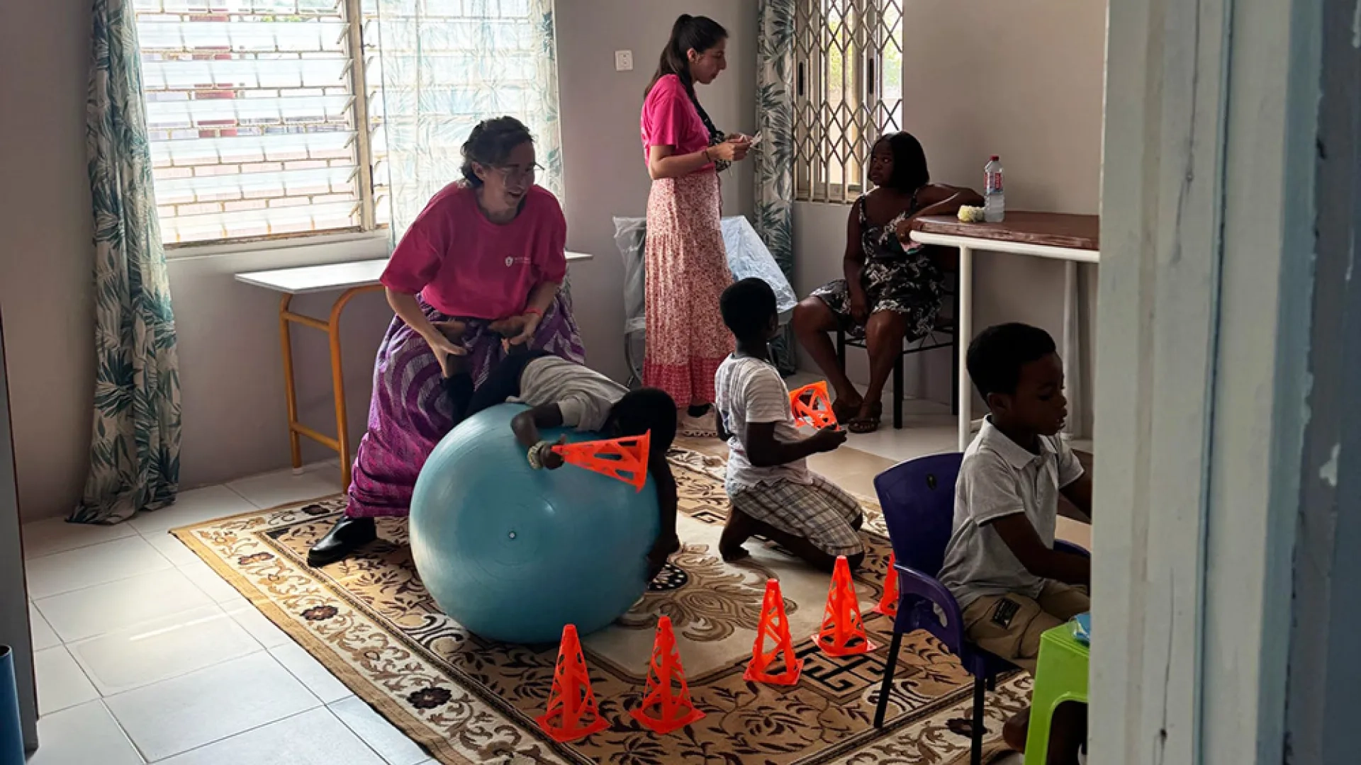 A child plays on the floor while another sits in a chair at a table while a woman in a chair watches them and another woman talks to a woman sititng in a chair