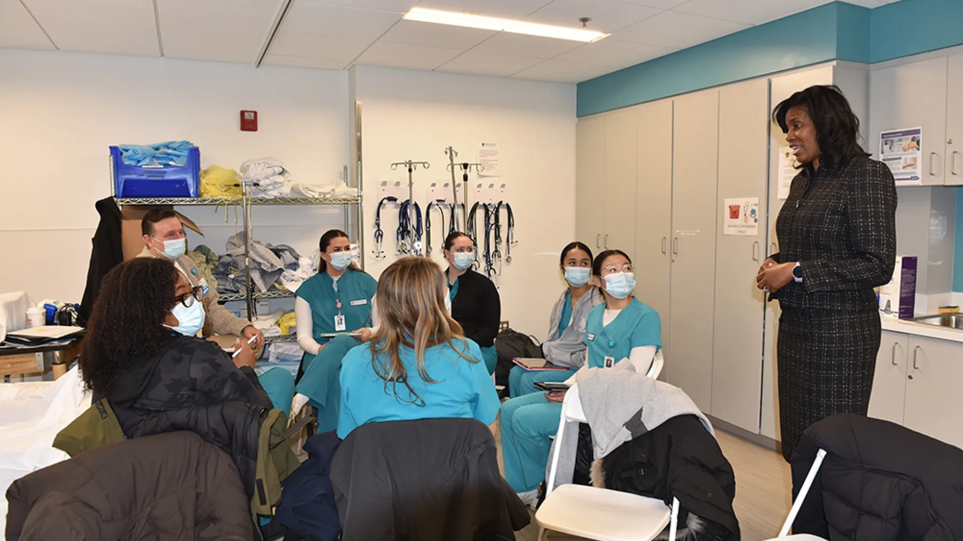 A woman stands talking to a group of people in medical scrubs sitting in front of her