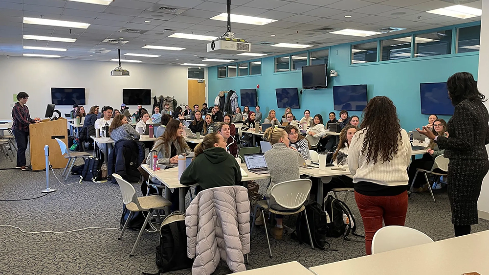 A classroom full of students listens to two people speaking at the front