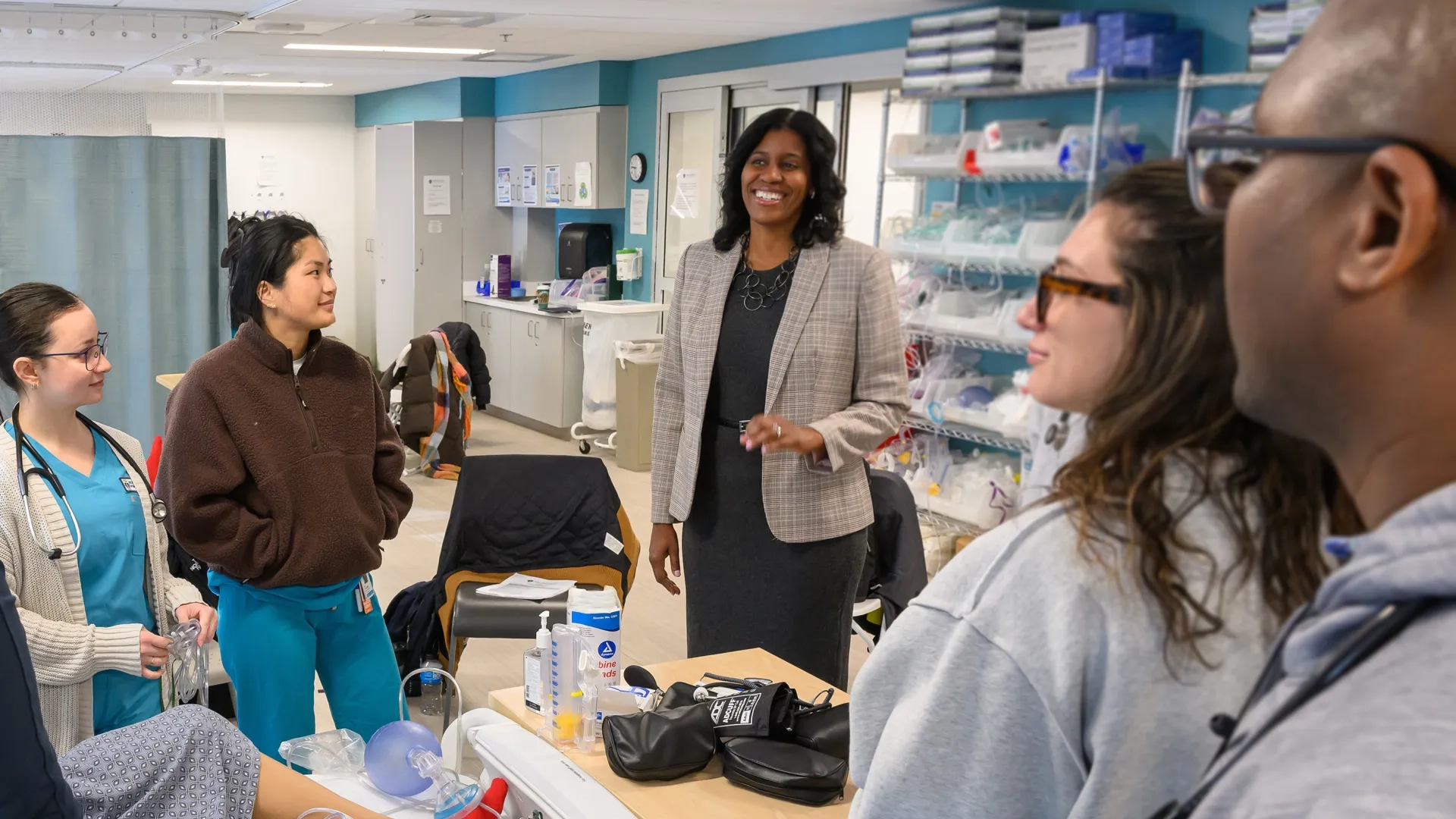 a woman speaks to nursing students in scrubs gathered around a hospital bed