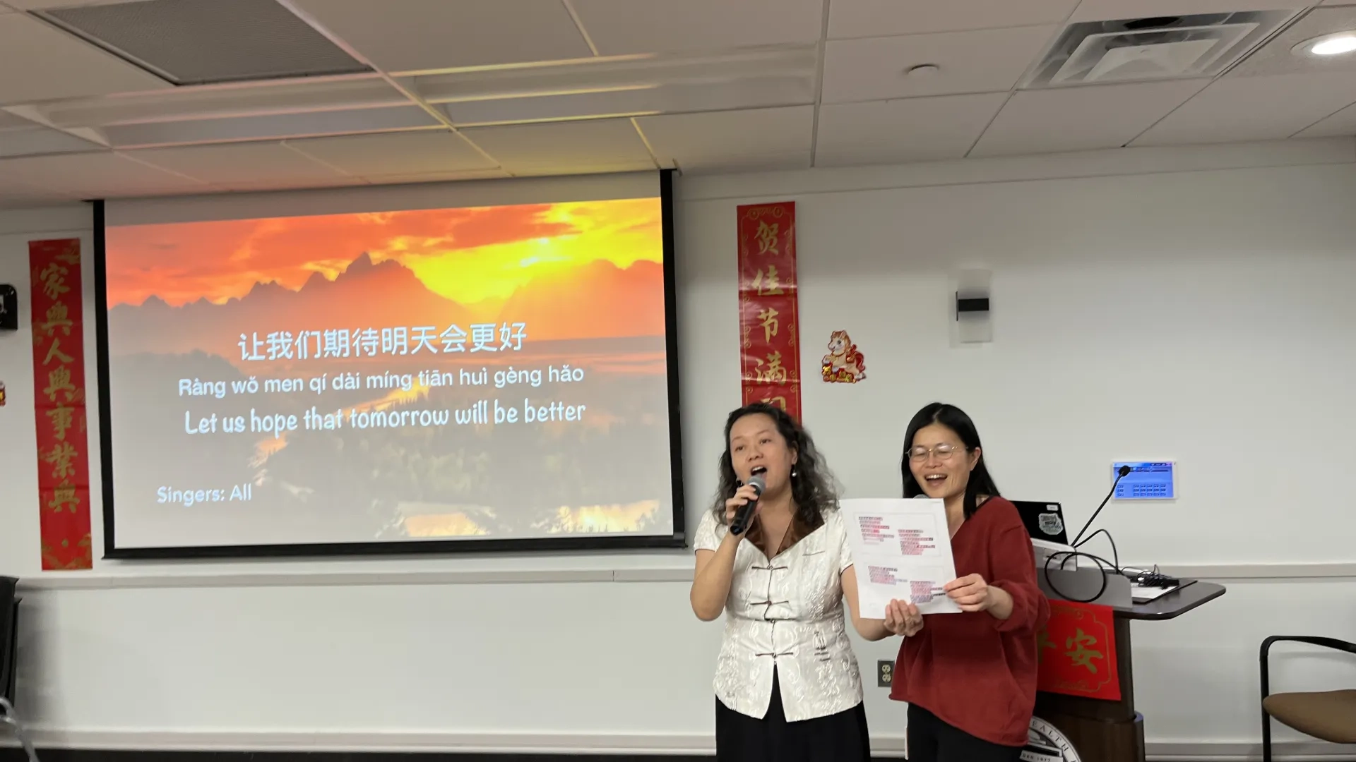 Two women sing in front of a screen with Chinese and English characters on it