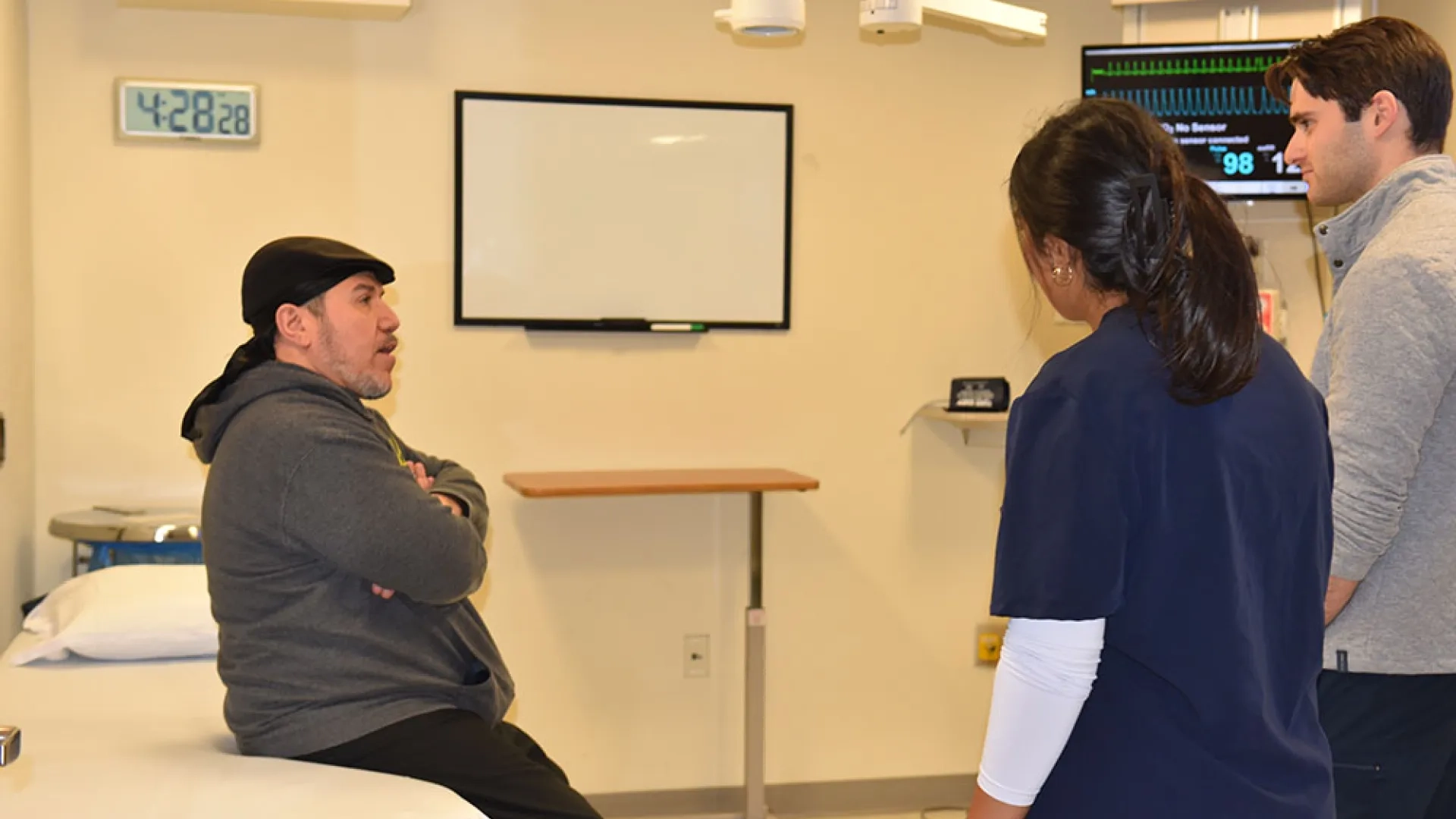 A man sits on a hospital bed talking to two healthcare professionals