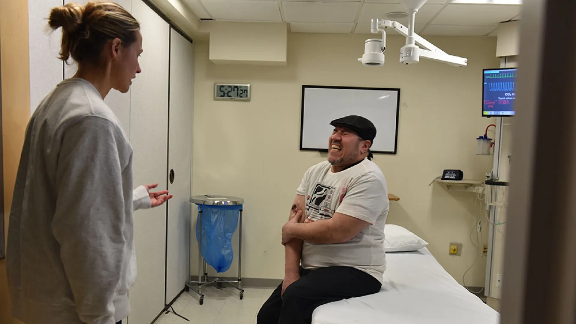 A man sits on a hospital bed holding his arm and talking to a medical professional