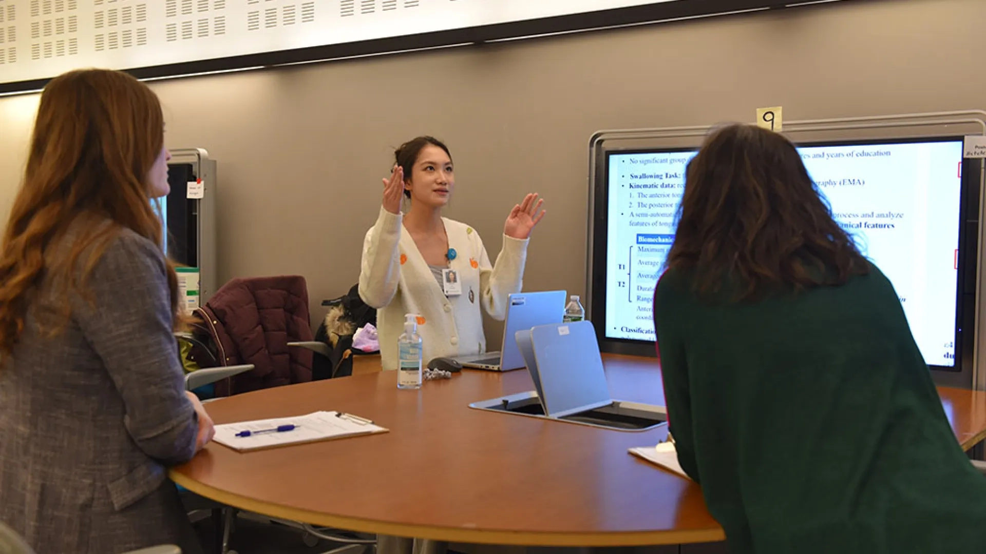 A woman stands in front of screen and talks to two people facing her