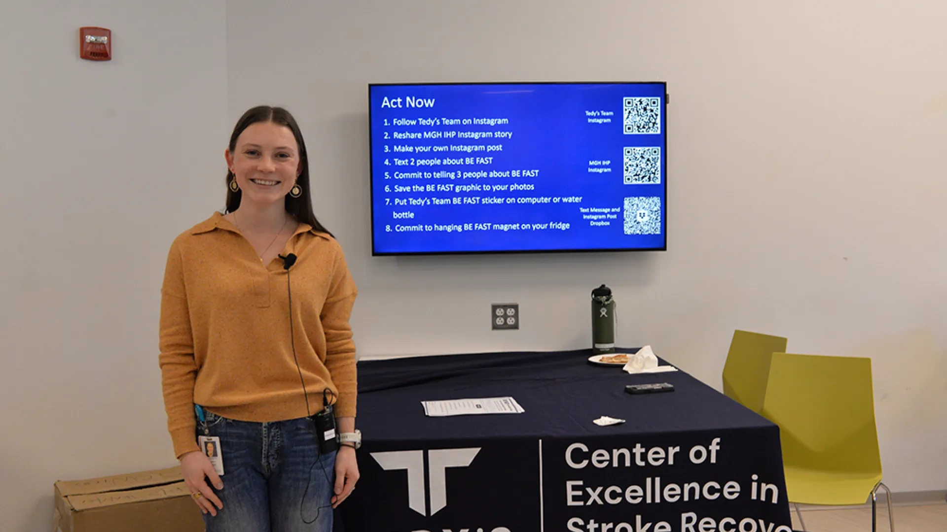 A woman stands in front of a table and screen 