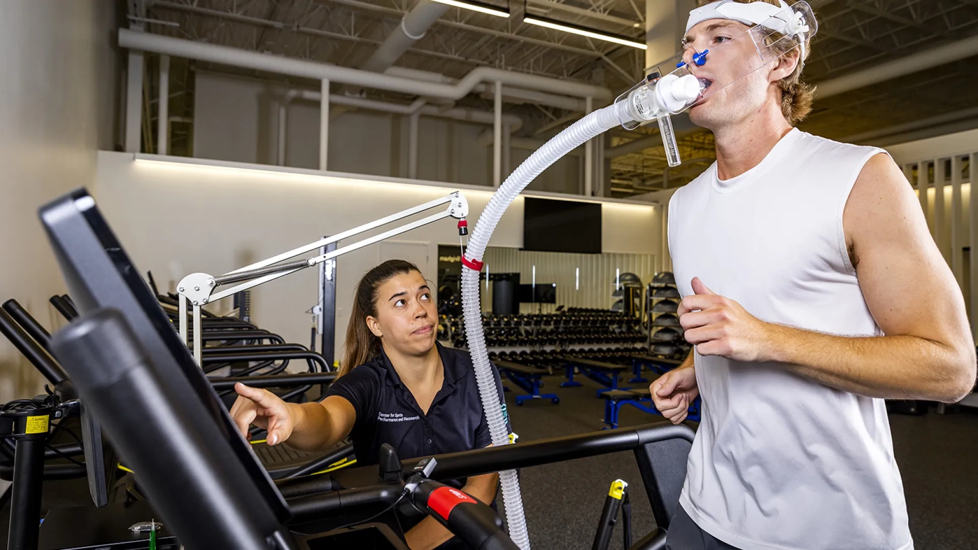 man runs on treadmill with a breathing tube in his mouth to measure output while a sports physical therapist looks on