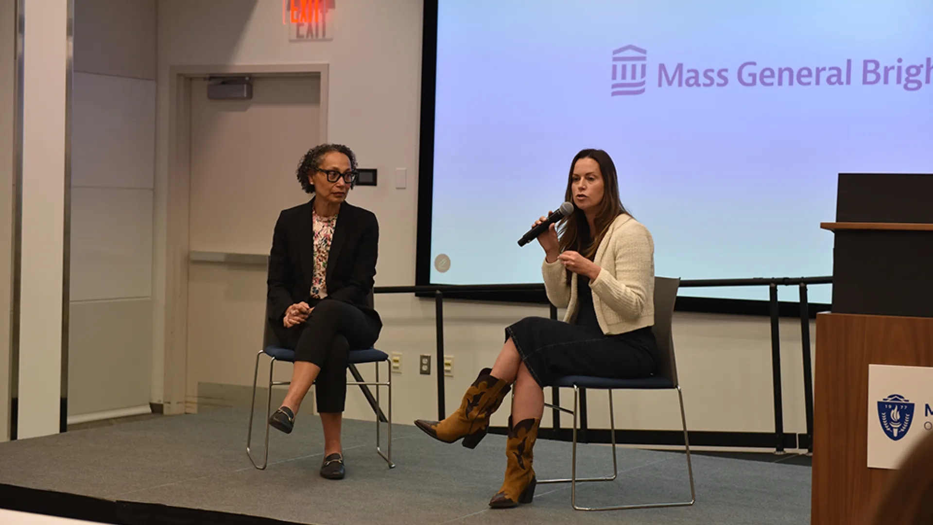 Two women sit on a stage with one speaking into a microphone