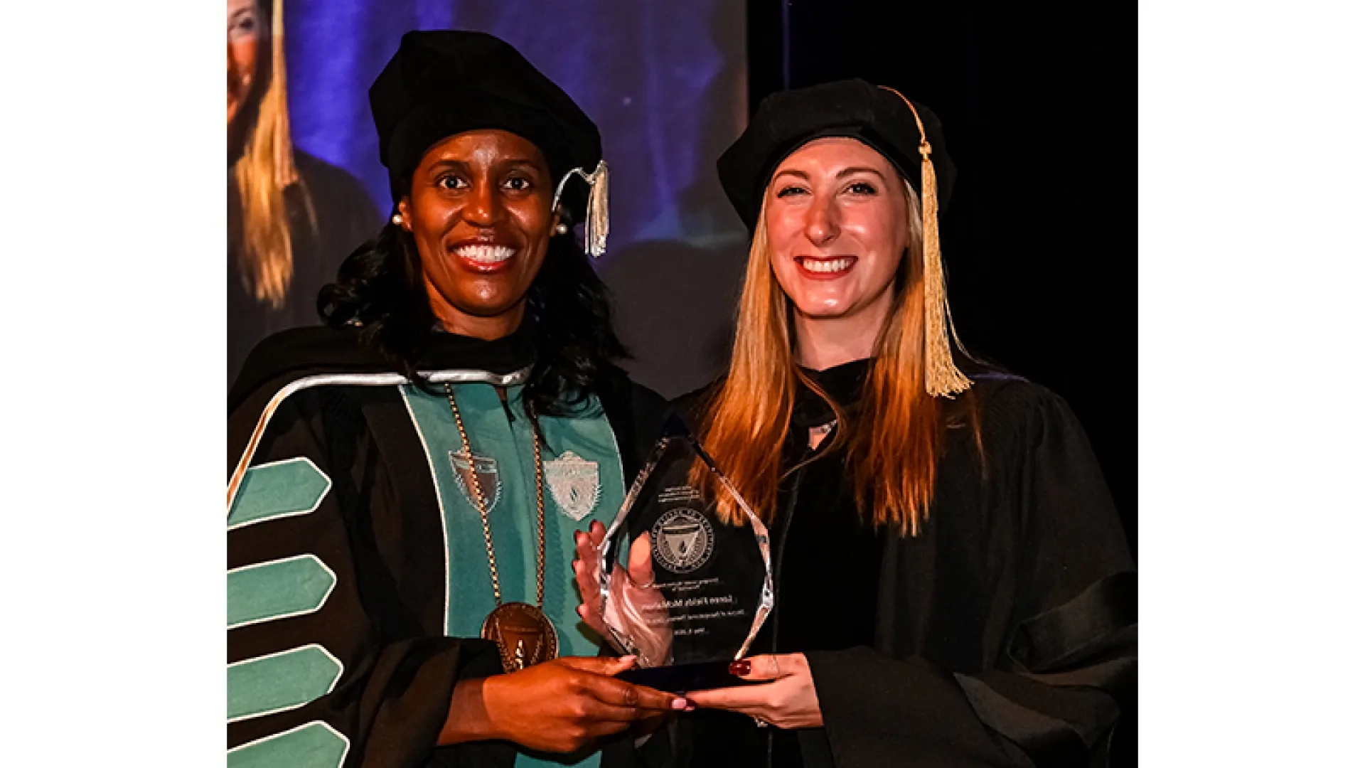Two women in graduation clothing holding an alumni award