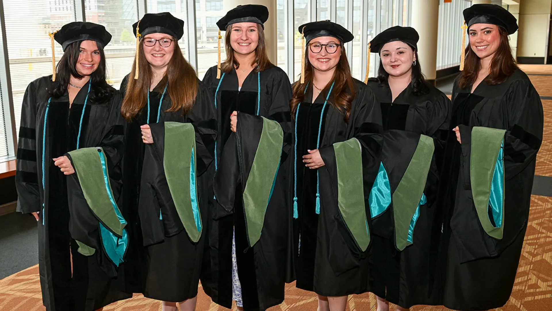 Six women wearing graduation gowns and holding academic hoods pose for the camera at commencement