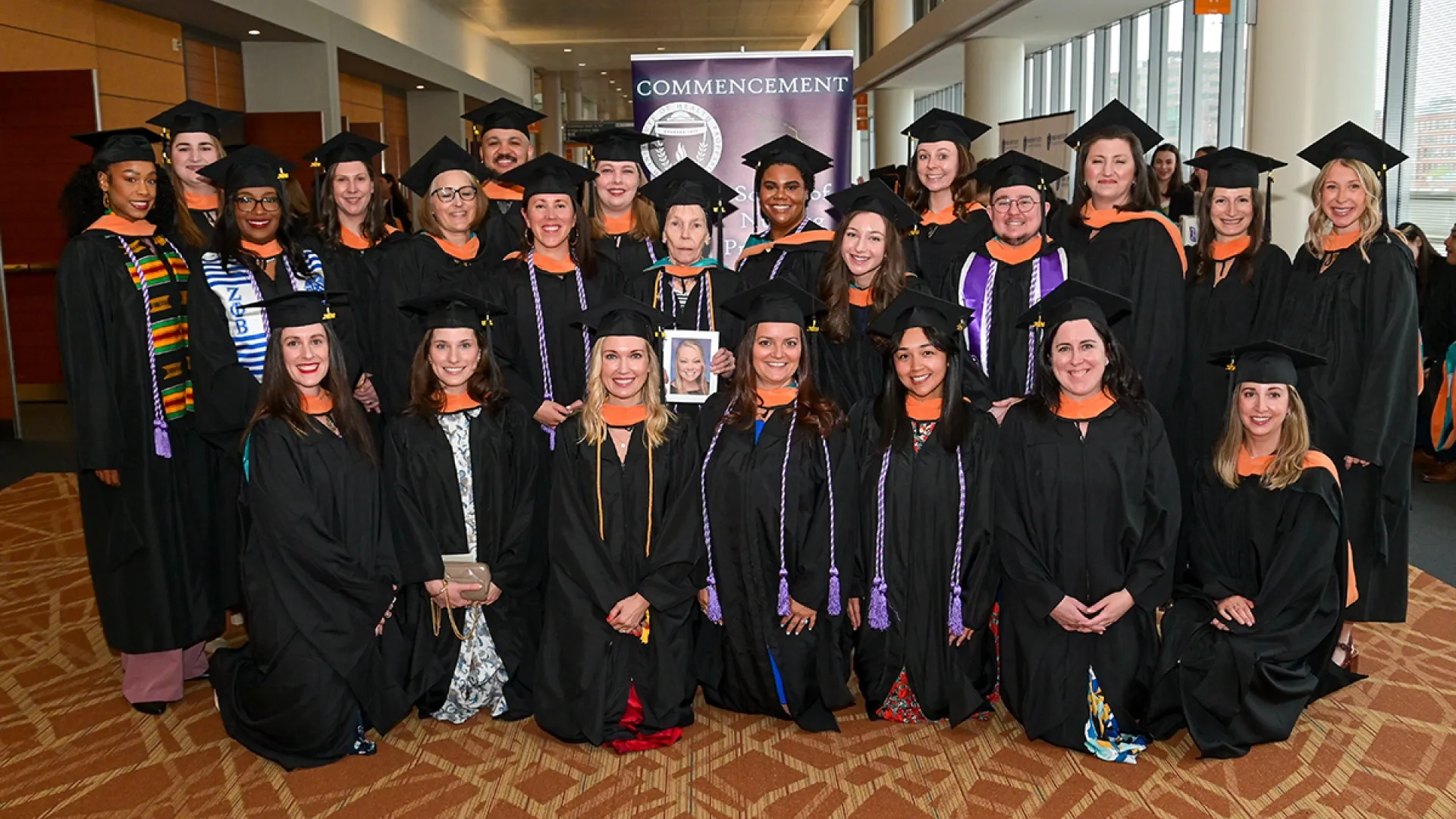 23 people wearing graduation gowns stand in three rows for a commencement photo