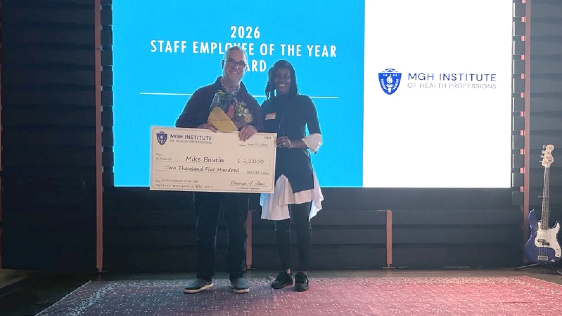 A man holding a big check stands next to a woman in front of a screen that says 2026 Staff Employee of the Year
