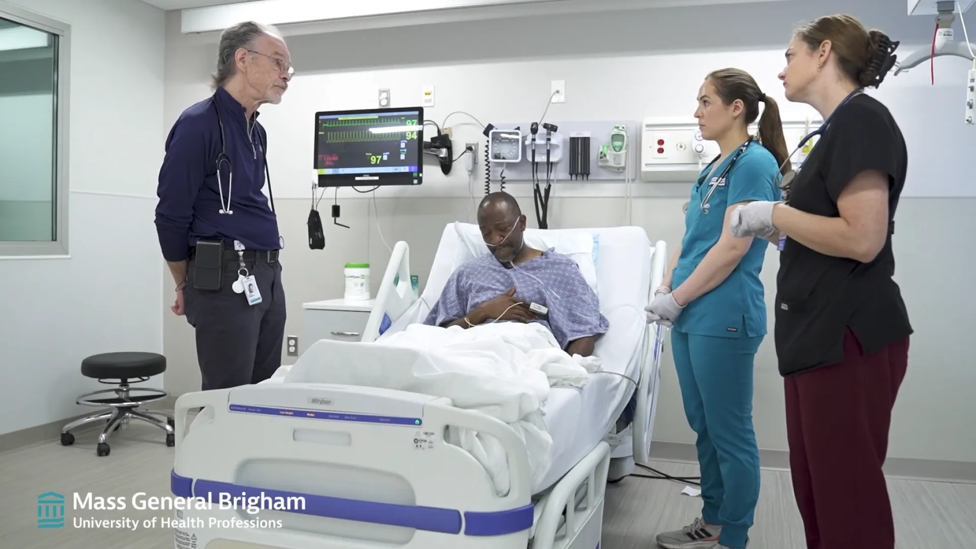 two women nurses in scrubs consult with a man in plain clothes across a hospital bed with a man in it with his hand on his chest