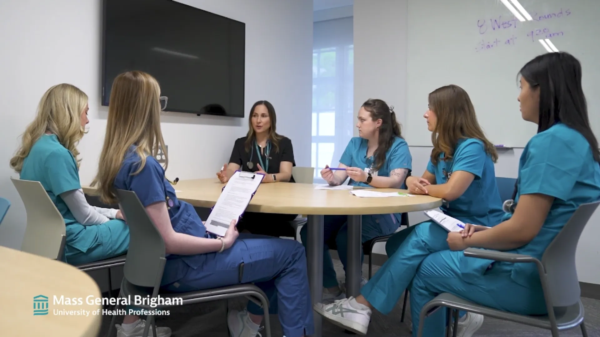 a group of students in scrubs sit around a table and discuss visits with their instructor 