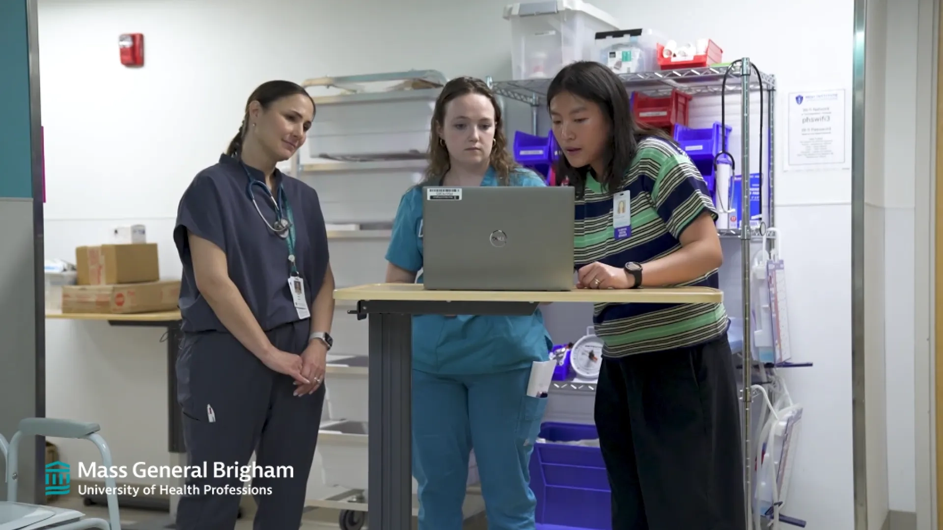 three women stand around a laptop in a hospital hallway, two wear scrubs one has on street clothes