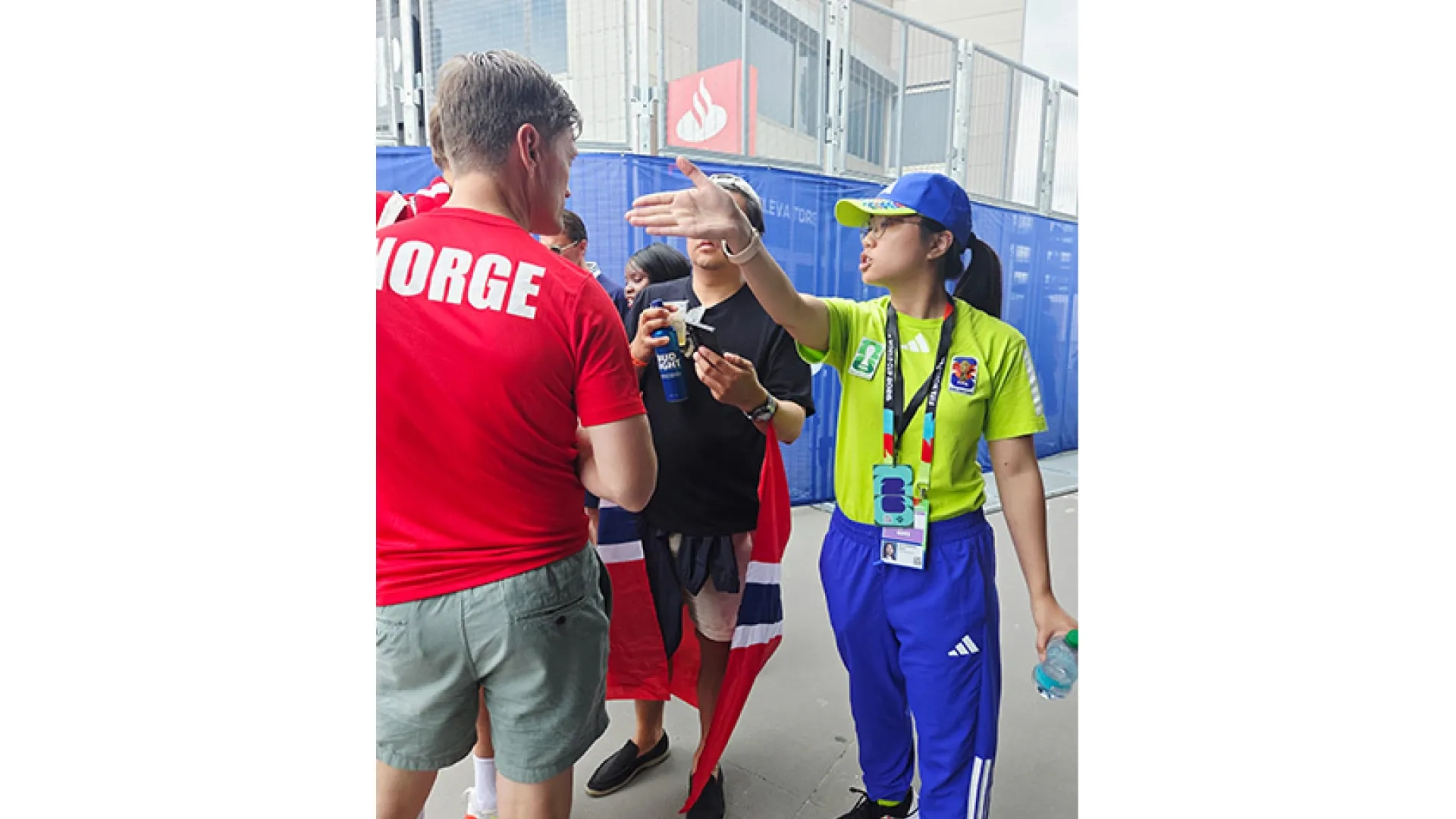 A woman wearing a yellow shirt and lanyard points in a direction for two people wearing items indicating they are from Norway