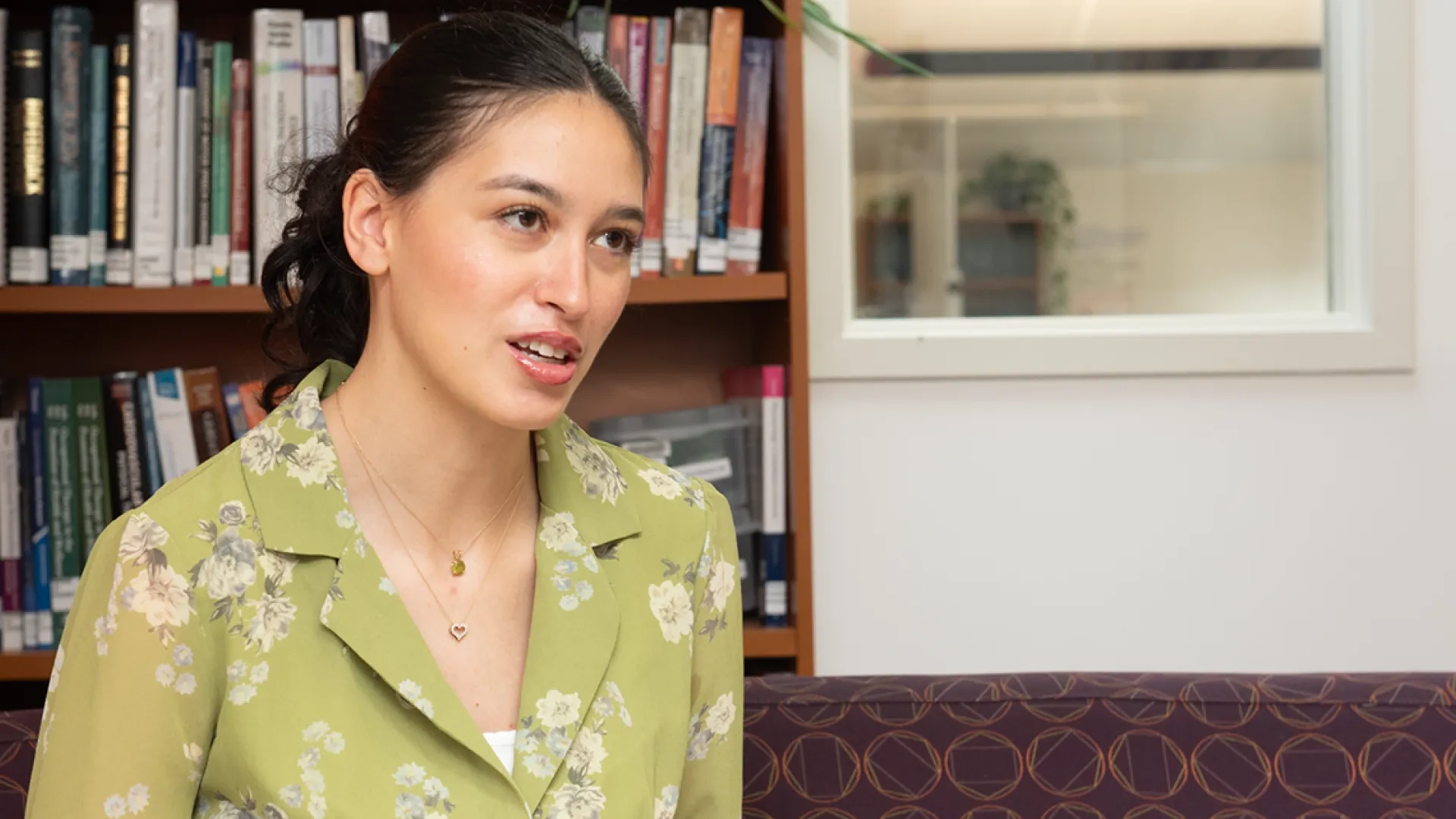 Nicole Vasquez sits on a couch with a bookcase behind her talking to someone off camera