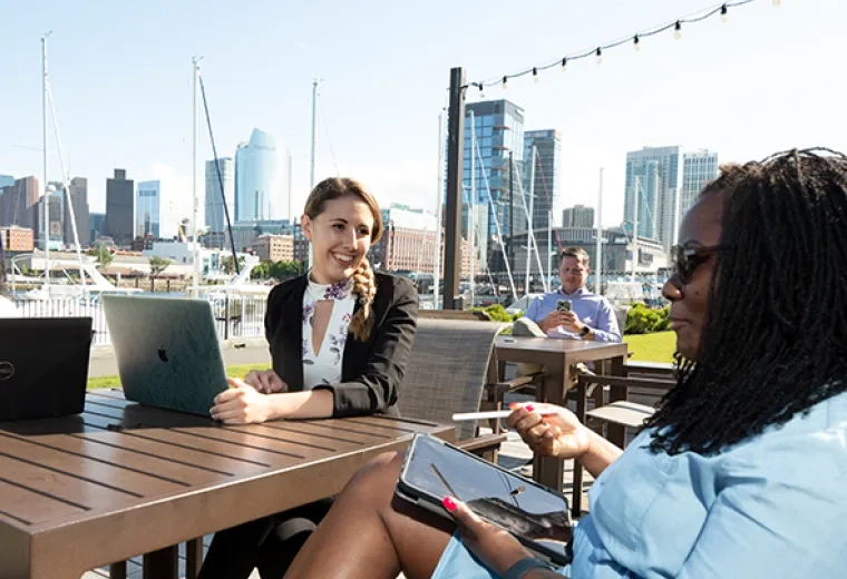 Woman in blazer speaks to woman in blue dress on a patio with boston skyline in the background
