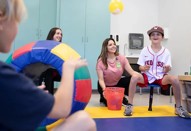 child in red sox jersey balancing on a stool smiles as a person in the foreground tosses a yellow ball to him