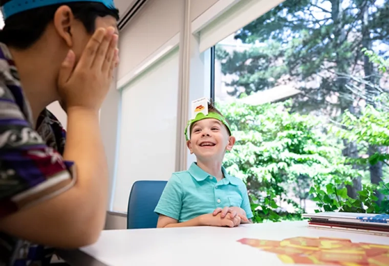 6 year old child smiles, sits at table playing a guessing game - a card on headband around his head says "i am spaghetti"
