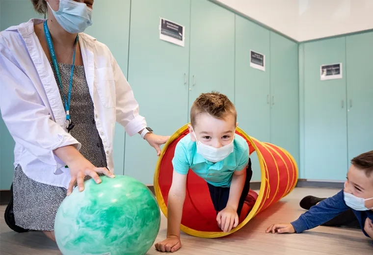 a little boy in a mask is helped by an IHP student to crawl through a cloth tube while his brother looks on