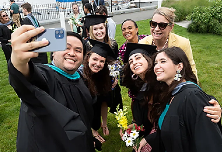 man and women in caps and gowns smile up at a cell phone 