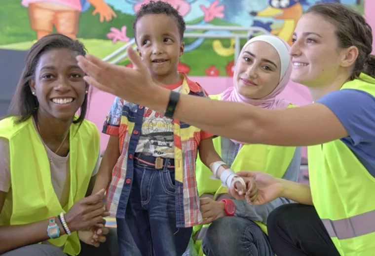 woman gestures to a little boy in a cast while two other women in yellow vests hold his hands