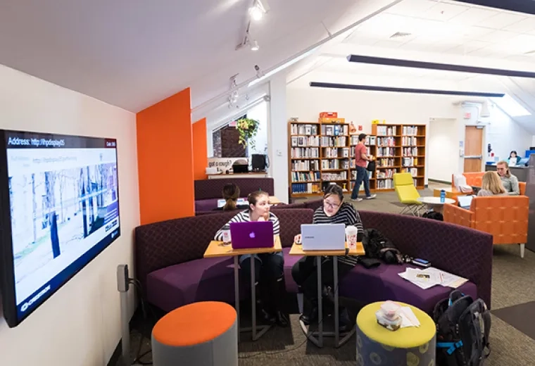 two women sit on a purple couch looking at laptops with a tall bookshelf in the background