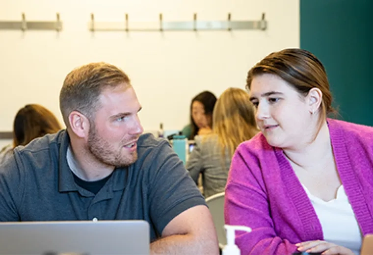 a woman leans over to look at a laptop while a man talks to her about something