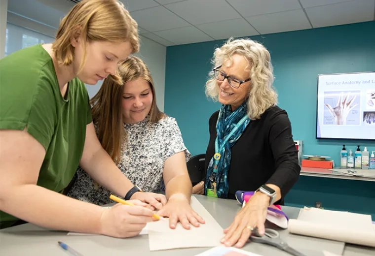 young woman with short strawberry blond hair traces another woman's hand on paper while another woman looks on