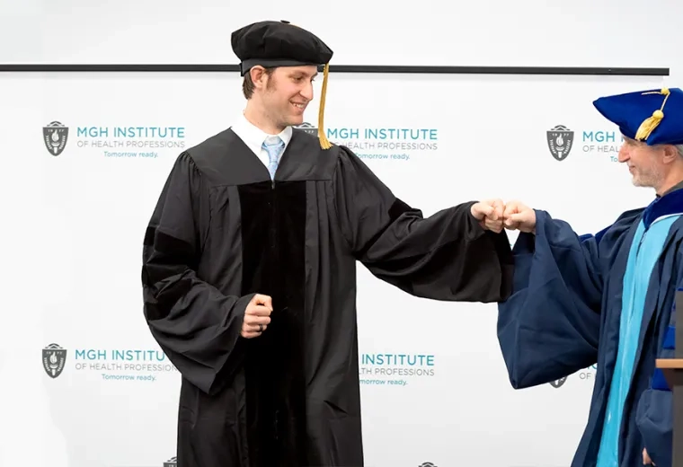 Man in graduation robe and cap fist bumps another man in blue graduation attire