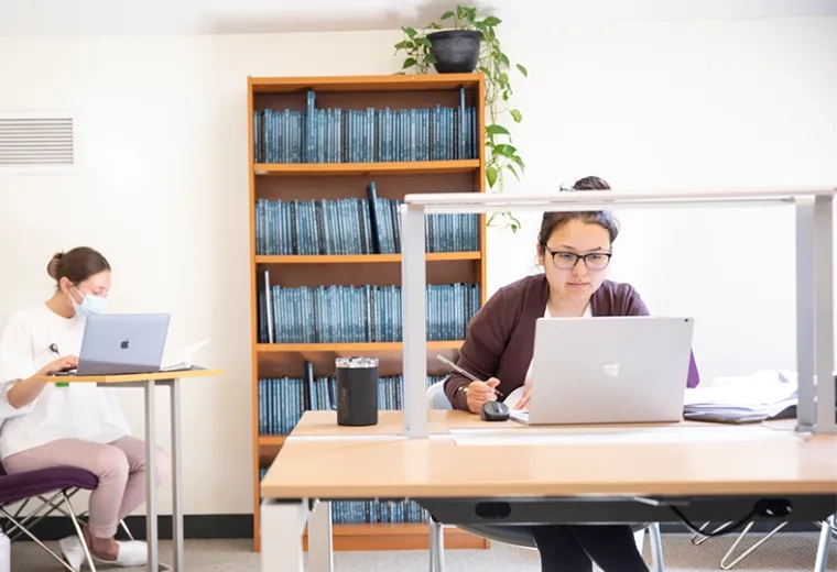woman sits at laptop in front of a shelf of books while another works on a laptop at a table next to her