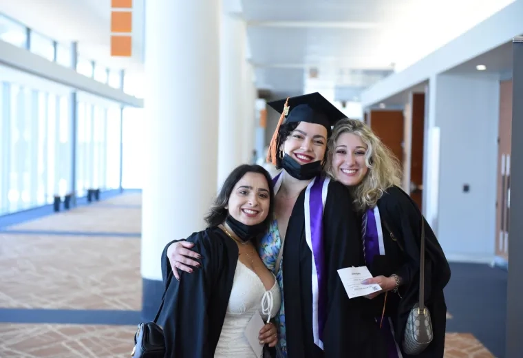 Three women in black graduation robes stand with their arms around each other smiling. One wears a graduation cap.