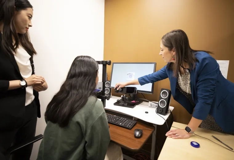 woman rests chin on black device and looks at a screen and camera while a woman points at the screen