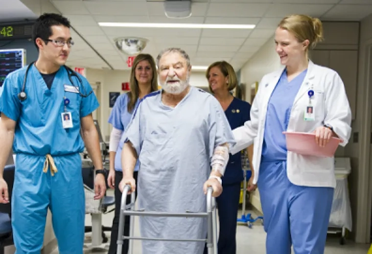 Four health professionals walk down a hallway with a patient whom is using a wwalker