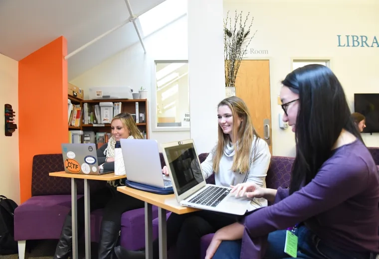 three women sit on a couch with laptops on tables