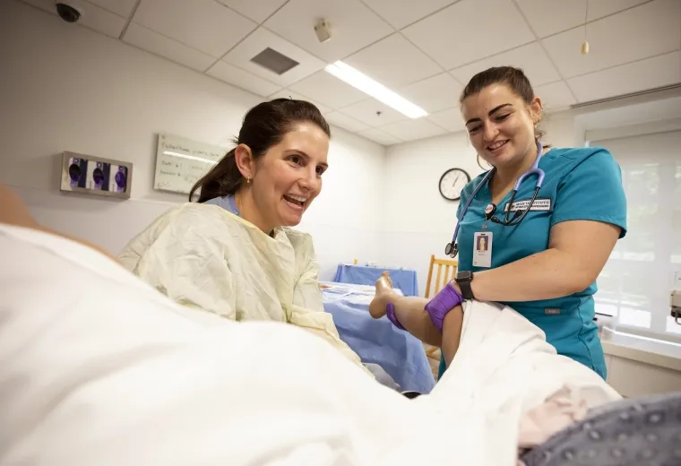 woman in scrubs holds a manikin leg while another women in a gown delivers a manikin baby