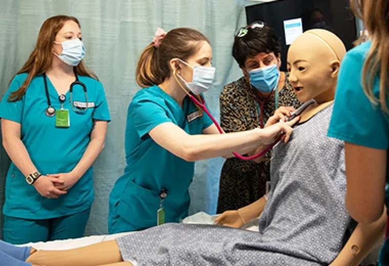 woman in scrubs uses stethoscope on a manikin while another woman in business attire helps position it