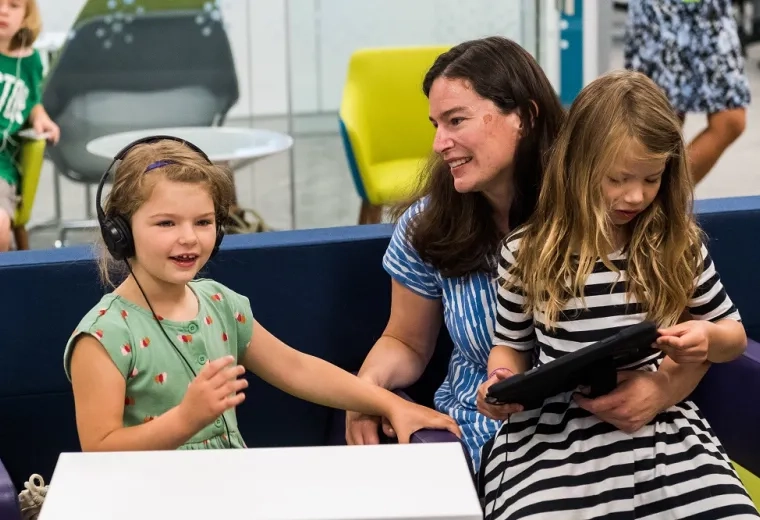 a little girl with headphones on sits next to a woman who has another little girl reading in her lap