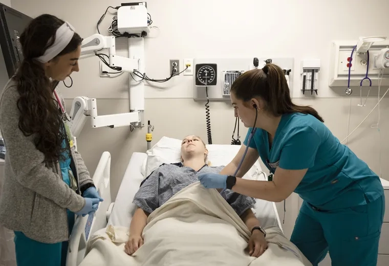Two professionals work over a patient in a hospital bed. One observes while the other holds a stethoscope to the patient