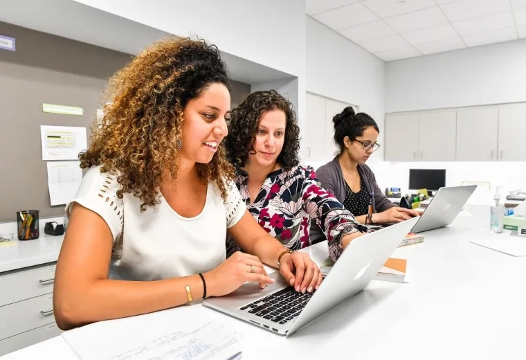 woman looks over shoulder of student at laptop