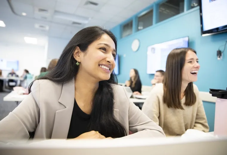 two females sit at a desk and laugh at something off camera