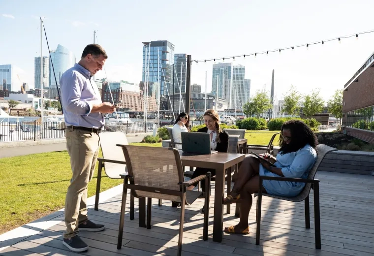 4 people on devices sit outside on a patio with the boston skyline behind them