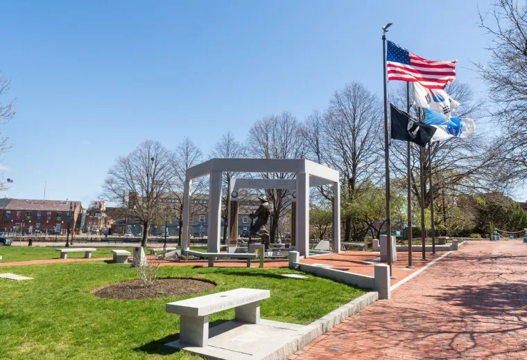 American Flag waves with POW MIA and other flags in front of a war memorial statue and a blue sky