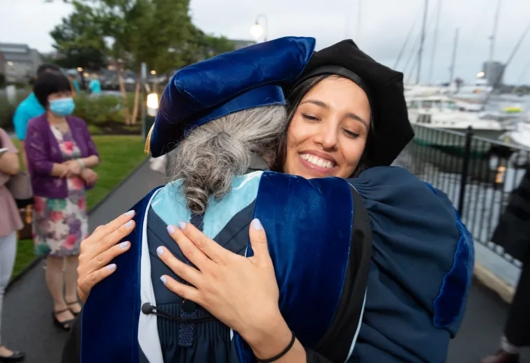 two people wearing dark blue graduation robes and hats hug happily 