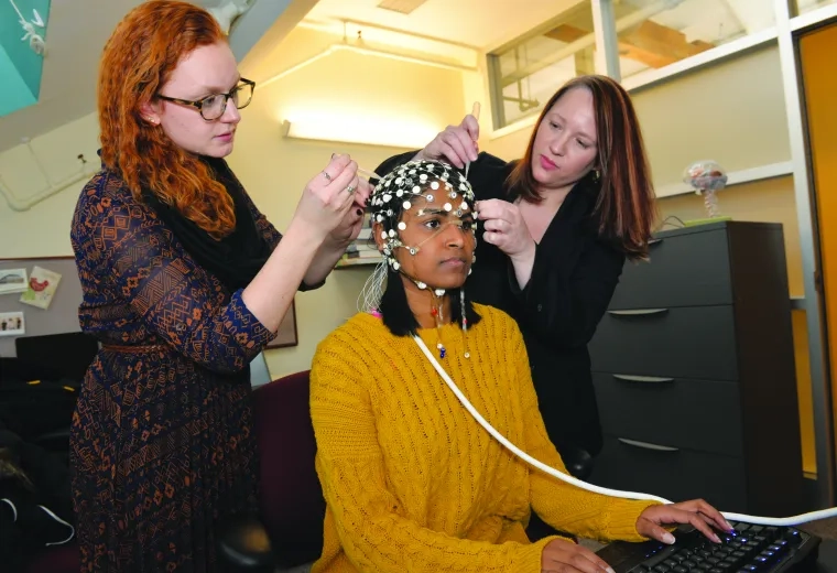 Two researchers place sensors on the head of a sitting person who is sitting and using a computer
