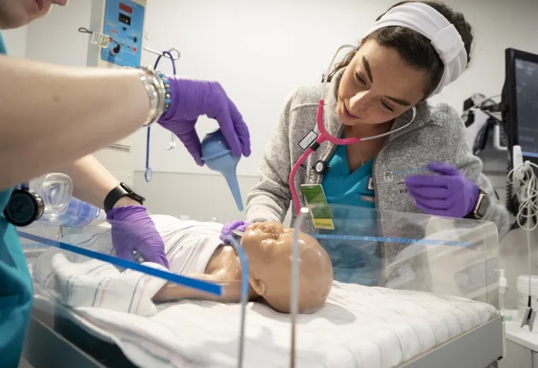 two women in scrubs look at a baby doll and use a suction bulb on it