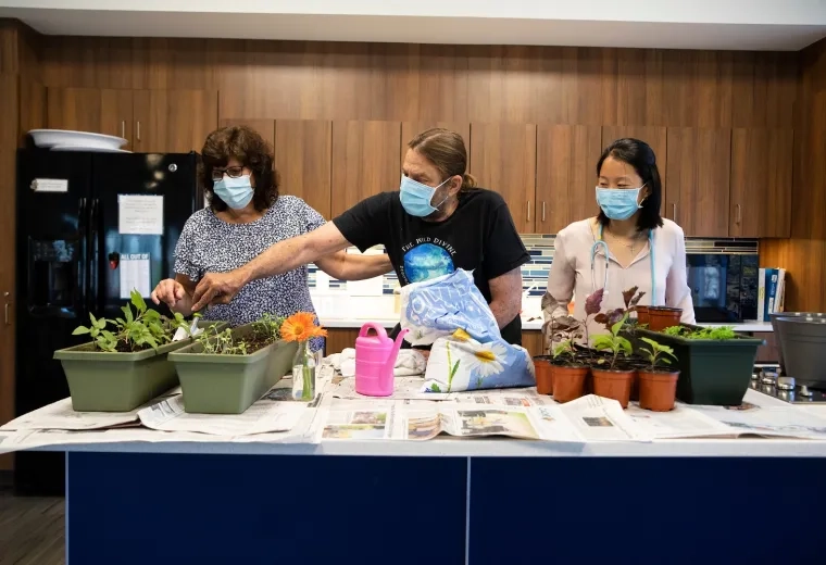 a man reaches across a table to dig in potted dirt while a woman steadies him